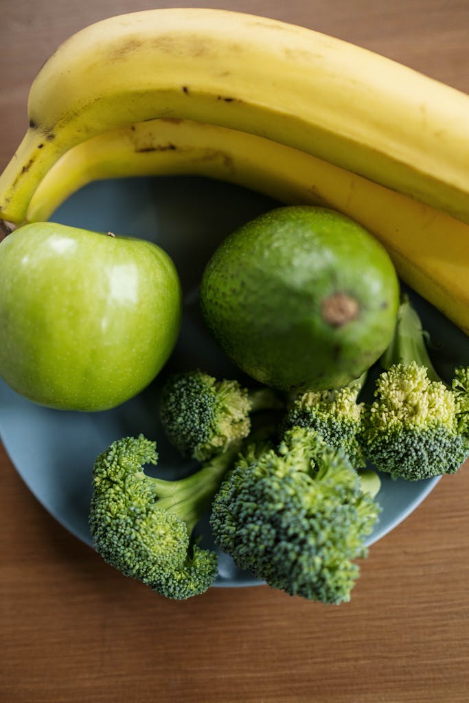A top view of fresh fruits and vegetables including bananas, apple, avocado, and broccoli in a blue bowl.